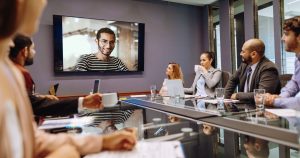 A team in a modern office setting attentively watching and interacting with a colleague on a large screen, indicating a virtual meeting in progress and highlighting the integration of remote participation with in-office work